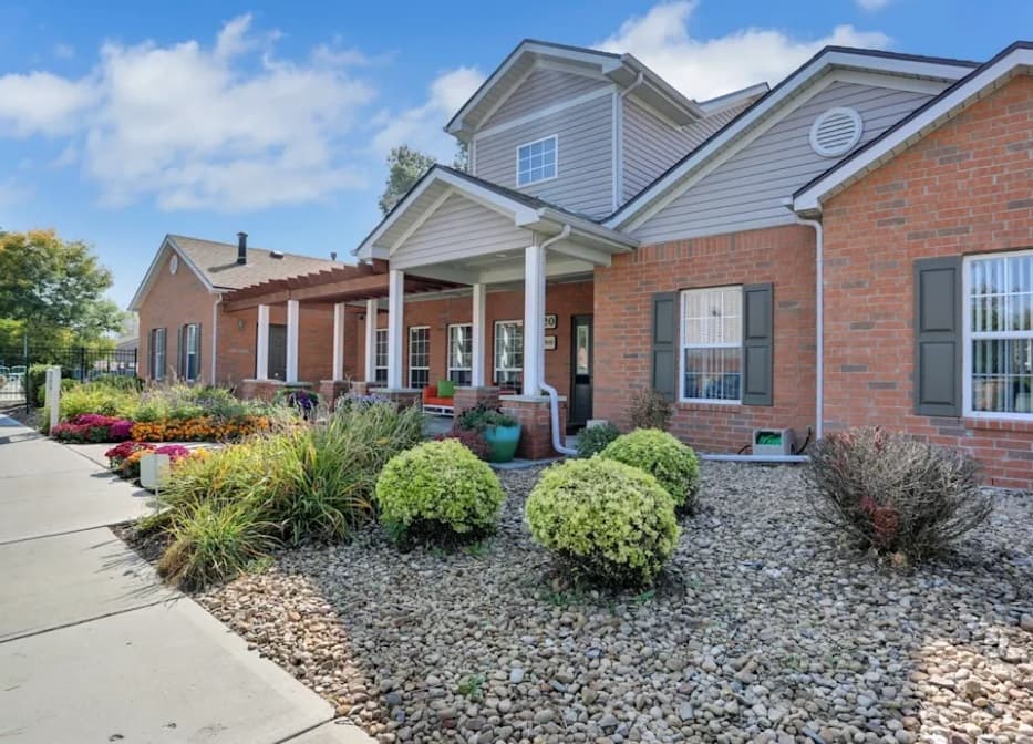 A brick building with white trim, large windows, and a covered porch at Bull Run Townhomes Fort Collins, surrounded by landscaped bushes, flowers, and decorative stone ground covering under a blue sky with scattered clouds.