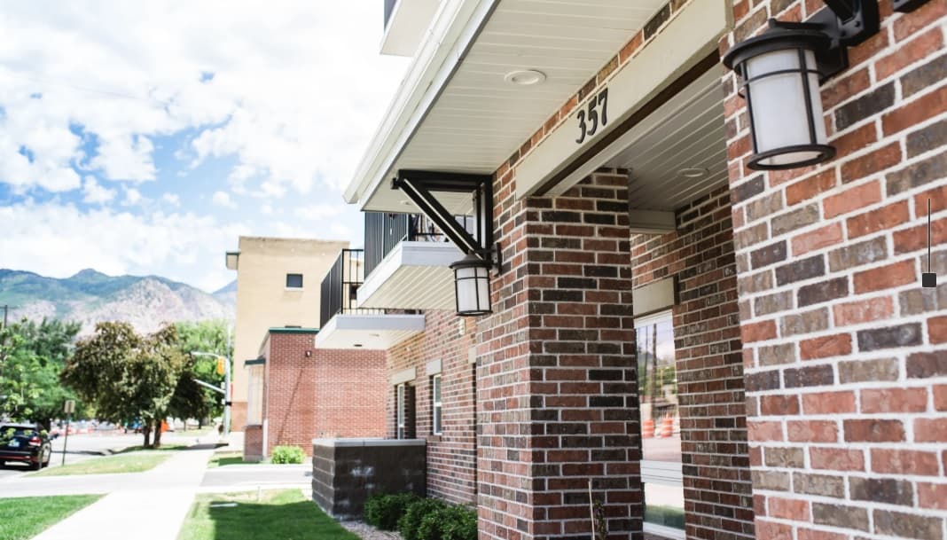 A row of modern brick apartment buildings with black trim and outdoor lights at City Garden Ogden, set against a sunny sky, green grass, sidewalk, and distant mountains in the background.