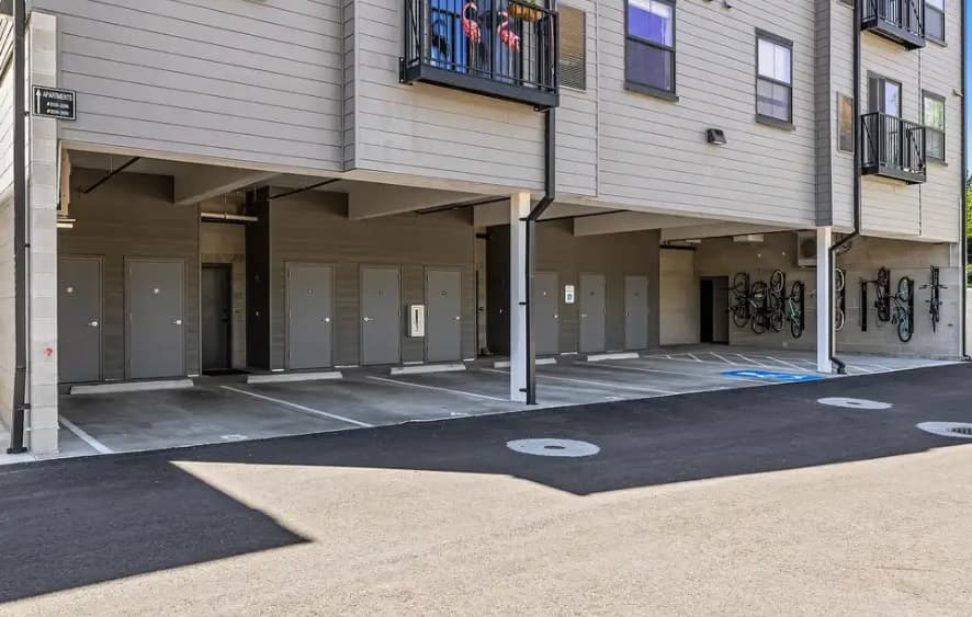 A row of covered parking spaces beneath 27th Street Crossing Boise, with locked storage doors behind each spot and bicycles mounted on racks to the right. One parking space features a blue handicapped symbol.