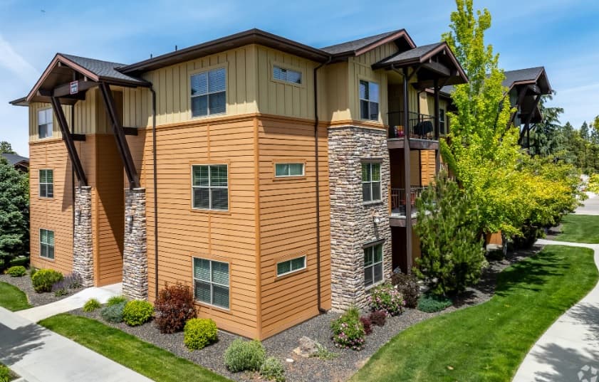 Retreat at Union Square Boise features a three-story apartment building with tan and brown siding, stone accents, and multiple windows. Enjoy well-maintained landscaping, green grass, bushes, and trees under a clear blue sky.