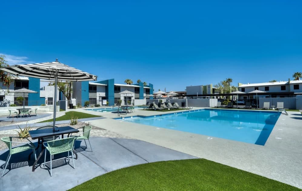 Modern Connect on Missouri Apartments Phoenix pool area with lounge chairs, tables, striped umbrellas, and green lawn under a clear blue sky. Multiple two-story buildings surround the refreshing pool.
