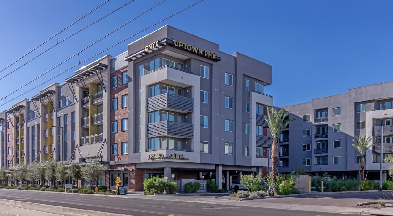 Modern apartment complex with multiple stories, balconies, and a sign reading Onyx Uptown PHX Apartments Phoenix; palm trees, greenery, and a clear blue sky complete the vibrant urban streetscape.