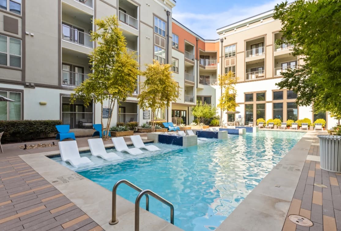Modern apartment complex, Rocklyn at Samuels Fort Worth, features a rectangular outdoor pool with white lounge chairs partially in the water, poolside seating, and trees surrounding the area. Multi-story buildings with balconies are seen in the background.