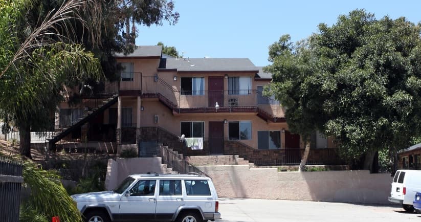 An apartment building with two stories features balconies, exterior staircases, and several doors. A white SUV and a white van are parked in front, with trees and greenery surrounding the area.