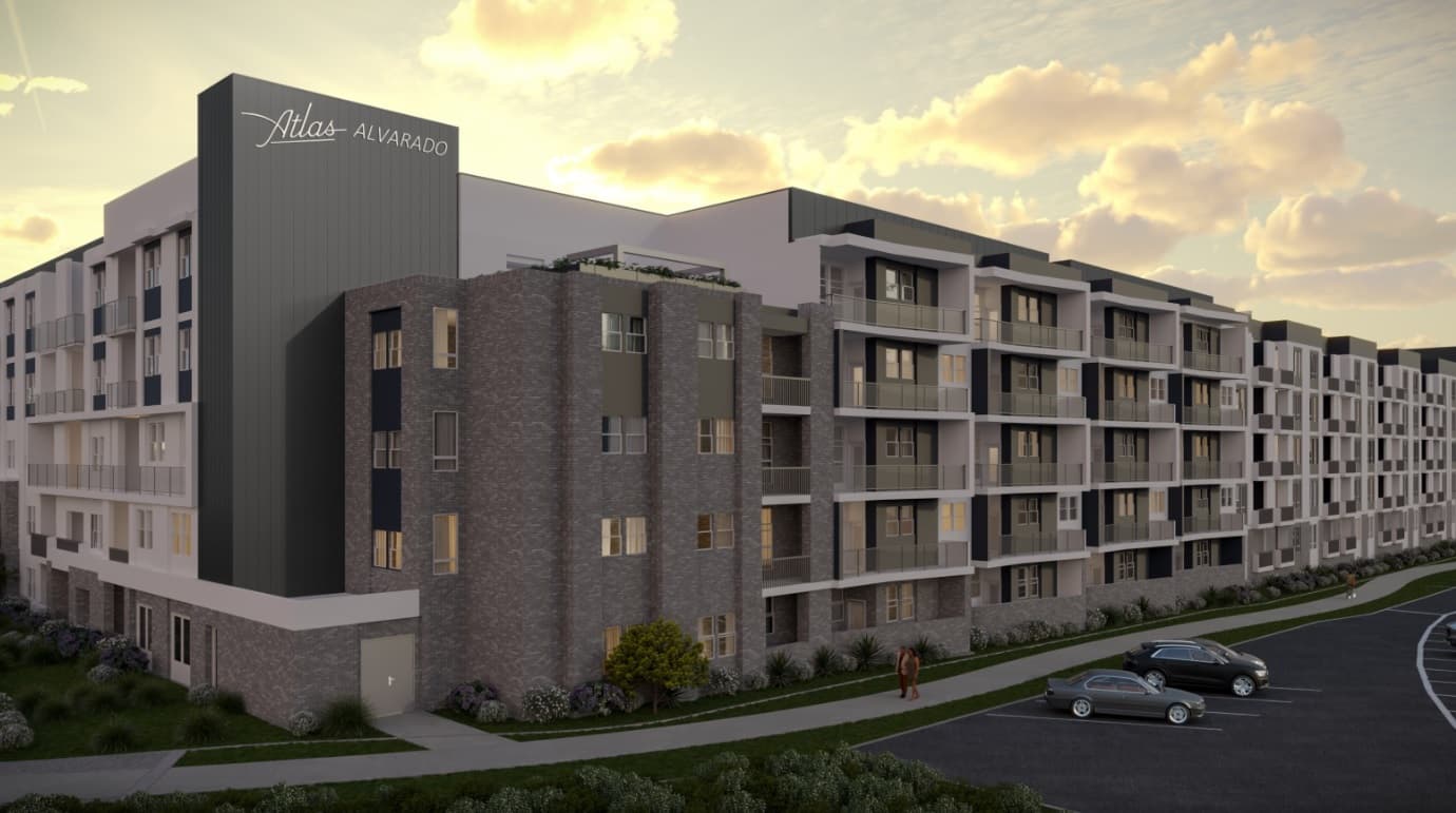 Modern mid-rise apartment building with balconies, named Atlas Alvarado, at sunset. Two parked cars and a person walking on the sidewalk are visible in front of the building, with landscaped greenery around.