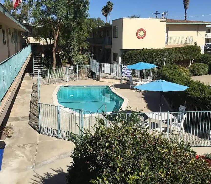 A small outdoor swimming pool surrounded by a metal fence sits in the courtyard of Villa Knolls Apartments, with lounge chairs and blue umbrellas nearby under a sunny sky.