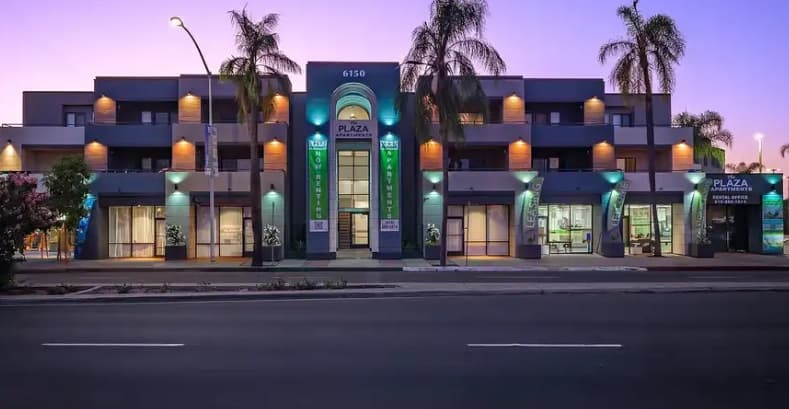 Modern two-story building with glowing Plaza Apartments signage and Dental Office at dusk, surrounded by palm trees and illuminated exterior lights, facing an empty street.