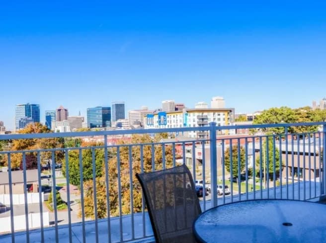 A glass patio table and chair sit on a balcony overlooking a city skyline, with tall buildings in the distance and a clear blue sky above. Trees with autumn foliage are visible below.