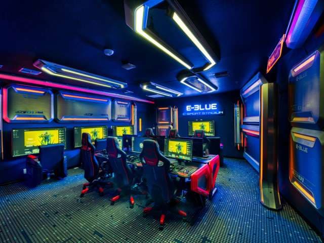 A futuristic gaming room with neon blue and orange lighting, multiple computer stations with large monitors, and ergonomic gaming chairs. A sign reads E-Blue Esport Stadium on the wall.