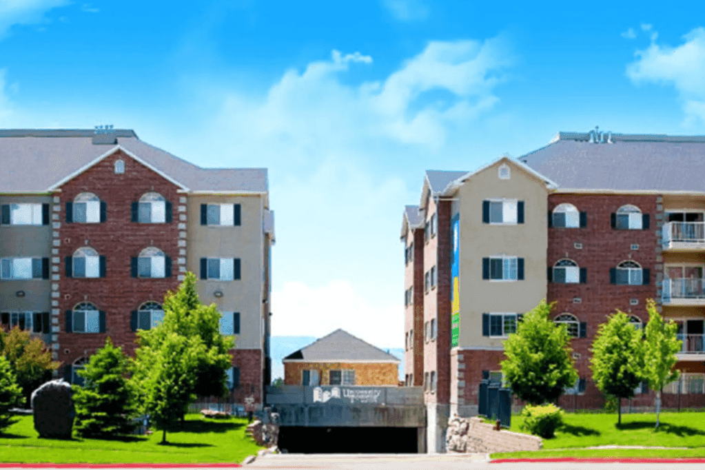 Two four-story apartment buildings at University Towers Orem, with brick and beige exteriors, stand on either side of a driveway surrounded by green trees and grass under a bright blue sky with scattered clouds.