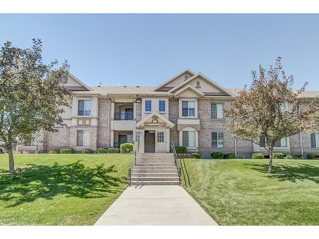 University Avenue Condos: A two-story apartment building with brick and stucco exterior, symmetrical windows, and a central entrance with stairs—designed by JosephThomas—surrounded by green lawn and trees under a clear blue sky.