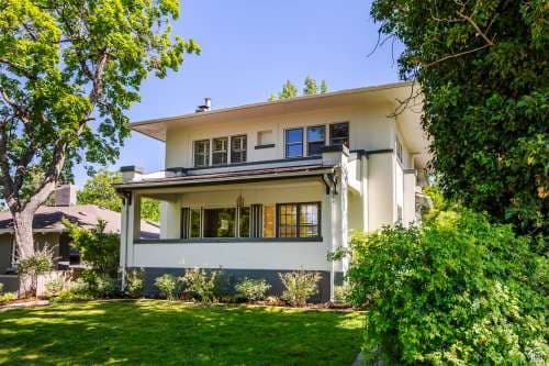 The Utah Prairie House in Salt Lake City is a two-story home with white and dark trim, large windows, and a covered front porch, surrounded by green shrubs, trees, and a well-kept lawn under a clear blue sky.