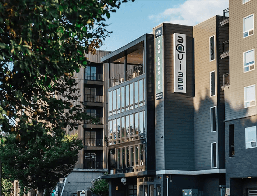 A modern multi-story apartment building with a sign reading aqui 353 and another vertical sign that says Now Leasing. Trees and blue sky are visible in the background.