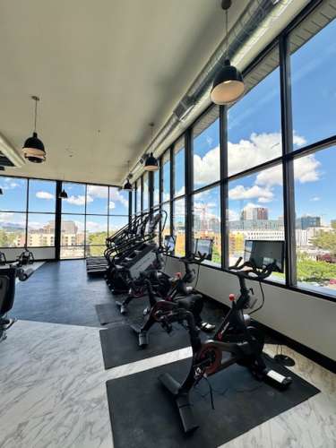 A modern gym at The Citizen Salt Lake City, with large windows overlooking a cityscape, features several stationary bikes and elliptical machines on a marble floor under hanging lights.
