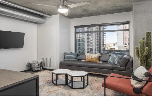 Modern living room at Atmosphere Tempe with a black sofa, colorful pillows, a red chair, large cactus plant, wall-mounted TV, and two hexagonal coffee tables. Expansive windows offer stunning views of city buildings outside.