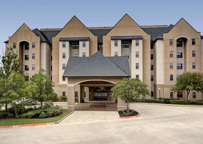A large beige multi-story hotel building resembling student housing with a covered entrance and peaked roofs, surrounded by trees, shrubs, and a paved driveway under a clear blue College Station sky—perfect for Callaway Villas residents.