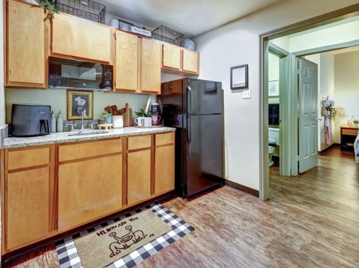 Cozy small kitchen with wooden cabinets, black appliances, and a playful dog doormat. Located in Callaway Villas student housing, this College Station home features wood-style flooring leading to a bedroom and bathroom.