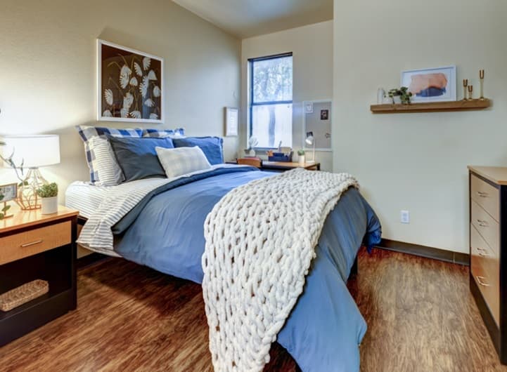 A cozy bedroom at Callaway Villas in College Station features a blue comforter, plaid pillows, a chunky white knit throw, wood floors, nightstands with lamps, a small desk by the window, and modern wall art—perfect student housing.