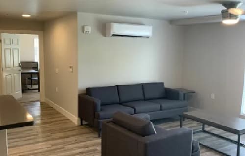 A modern living room with light-colored walls, wood flooring, a dark gray sofa and armchair, a coffee table, and a wall-mounted air conditioner. An open doorway leads to another room with a desk and chair visible.