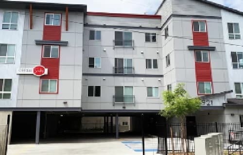 Coffee House Apartments is a modern four-story building with gray and white siding and red accents. The ground level offers open parking, and a small tree sits near the entrance. Several windows and balconies enhance the building’s inviting look.