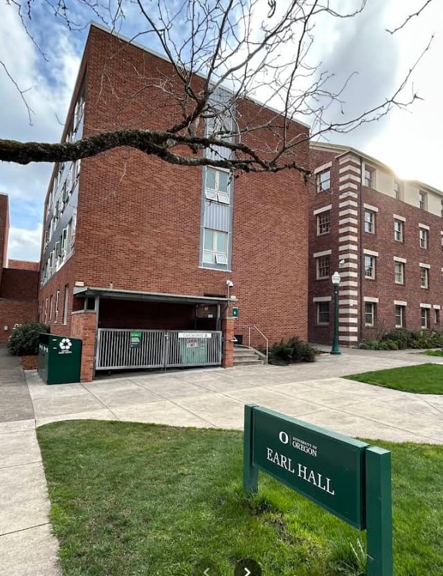 A red brick dormitory building with multiple windows and a green sign in front reading University of Oregon Earl Hall, set on a sidewalk and grassy area under a cloudy sky.