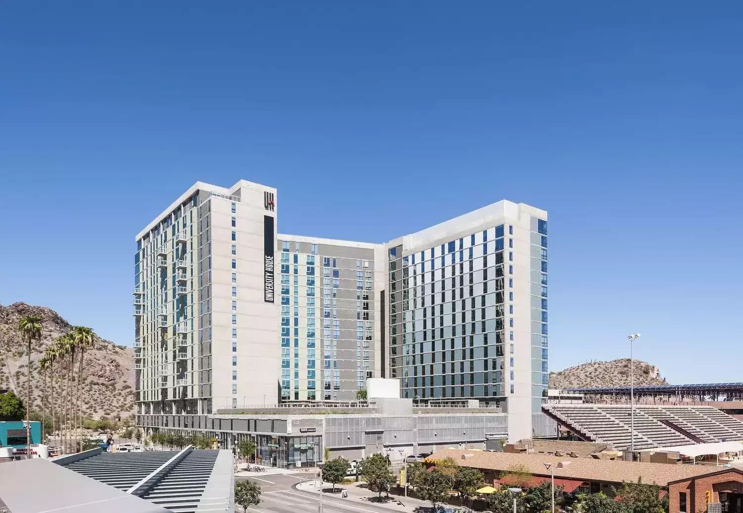 A modern high-rise hotel with glass windows stands against a clear blue sky near University House Tempe, with a mountain and palm trees in the background and a sports stadium visible to the right.
