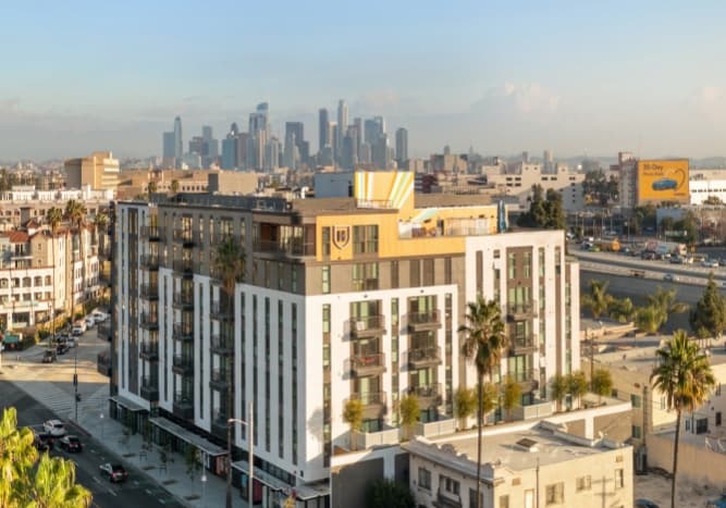 A modern mid-rise apartment building with balconies stands in the foreground, set against an urban cityscape near the Los Angeles Coliseum, with palm trees and downtown skyscrapers in the background under a partly cloudy sky.