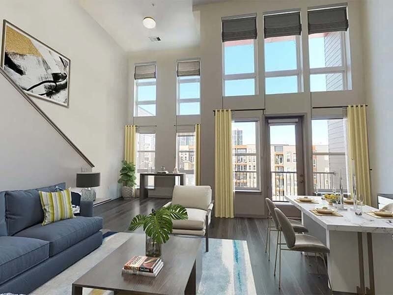 Bright, modern loft apartment with high ceilings, tall windows, gray sofa, armchair, dining table set for four, indoor plants, wall art, and city views of Viceroy Observatory Park through large windows with yellow curtains.