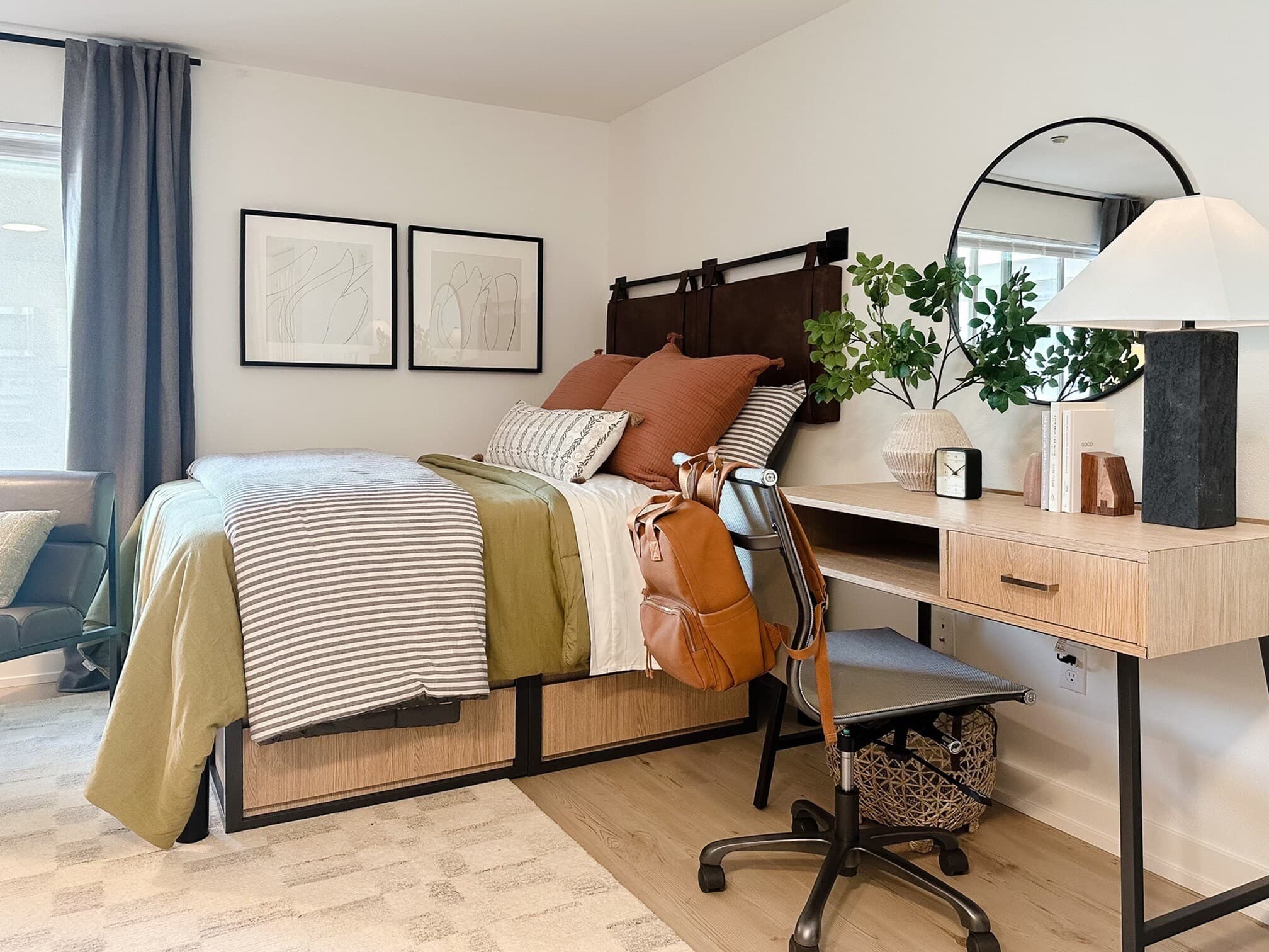 A modern, cozy bedroom in Yugo Seattle student housing with a neatly made bed featuring green, white, and brown bedding, a wooden desk with a lamp, books, potted plants, a chair, and a brown backpack. Minimalist art hangs above the bed.