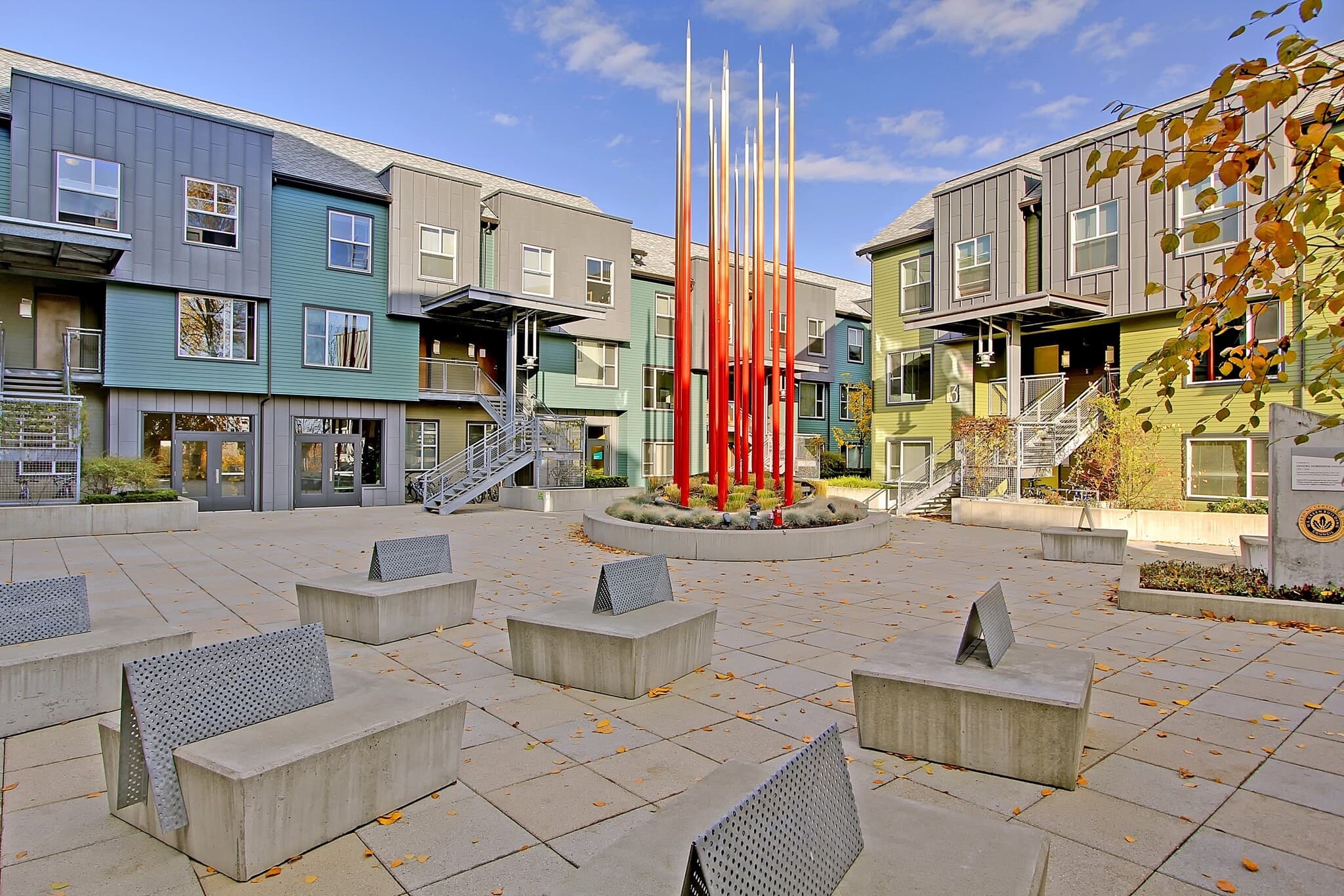 A modern courtyard at Nordheim Court features abstract metal benches, a circular planter with tall red poles in the center, and colorful UW Student Apartments surrounding the open space under a blue sky.