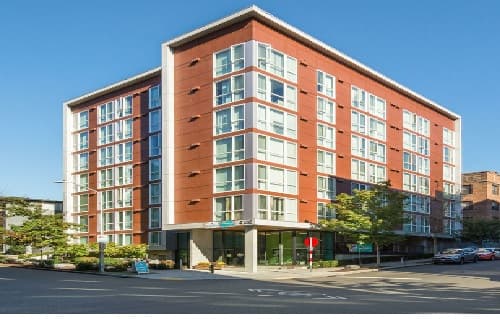Nora Apartments is a modern mid-rise building with large windows and a red and white exterior, standing on a sunny street corner with trees and clear blue sky in the background.