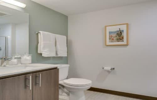 Modern bathroom at Nora Apartments featuring a toilet, wooden vanity, towel rack with two white towels, glass jars on the counter, and framed artwork above the toilet paper holder.