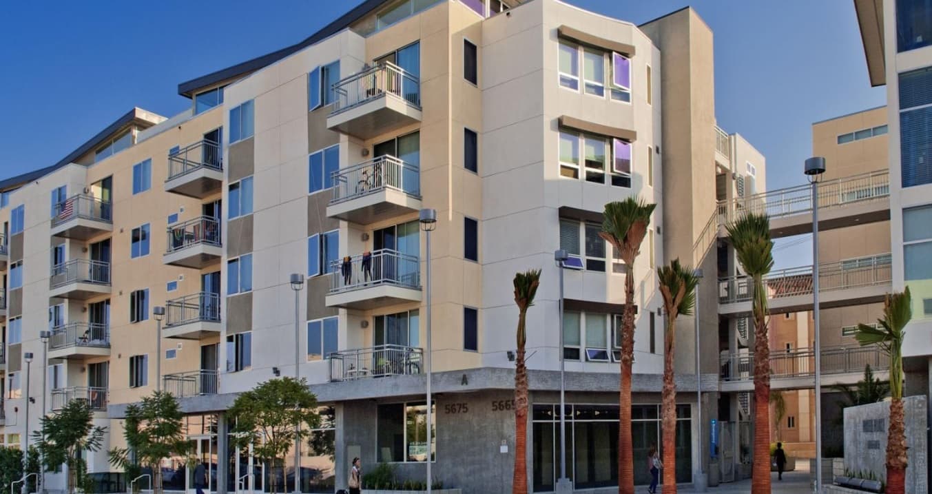 Modern five-story apartment building at Paseo Place San Diego with balconies, large windows, and a ground-floor retail space. Several palm trees and small shrubs line the sidewalk in front. The sky is clear and blue.