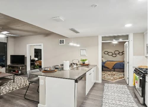 Modern open-concept kitchen with a large island and stainless steel appliances at The Standard at Seattle; living area features a TV and gray sofa; bedroom with a patterned rug and blue bedding visible through open doors.