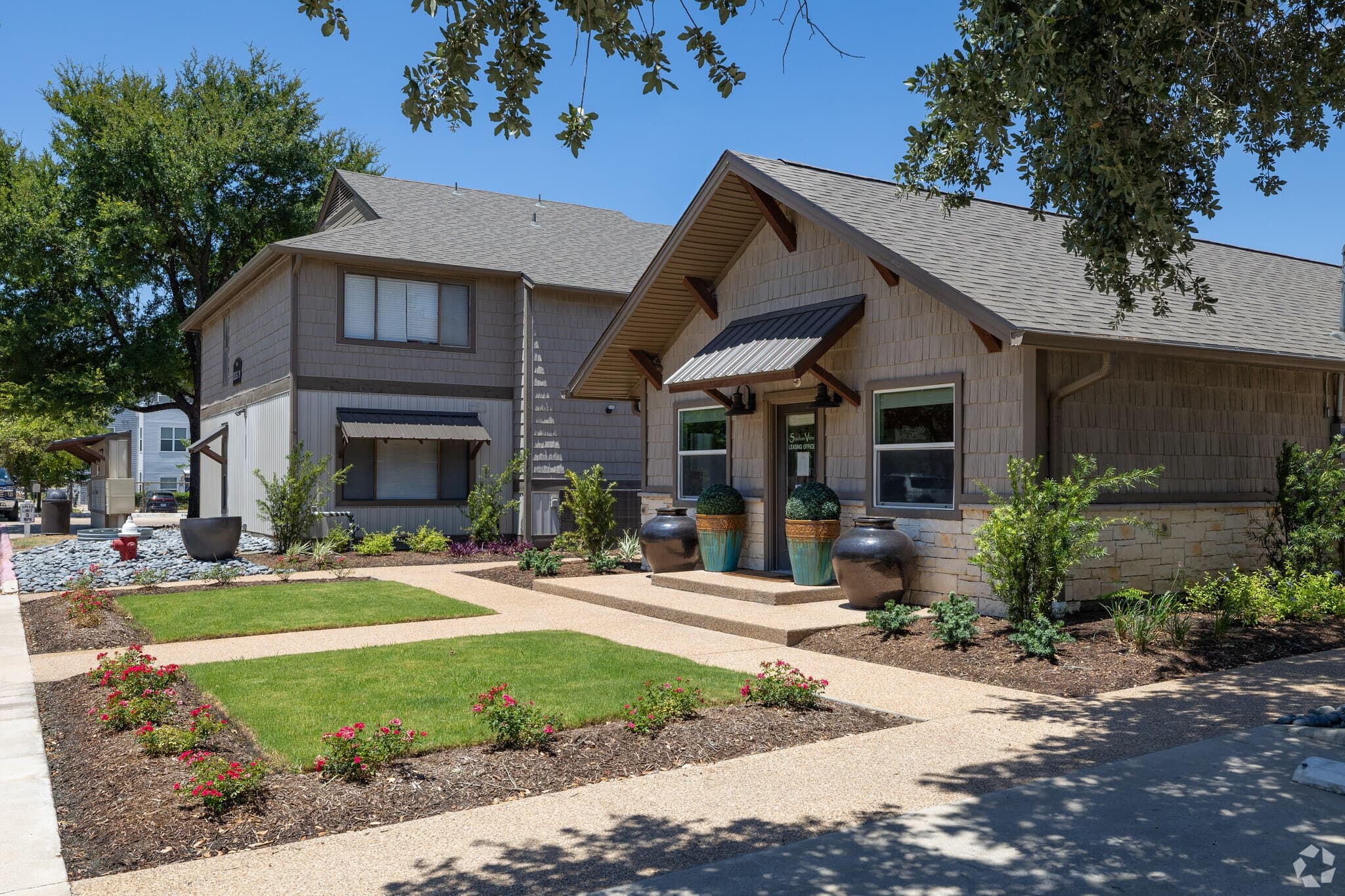 A modern two-story house and a single-story house sit side by side near Stadium View Apartments, surrounded by landscaped gardens with green lawns, shrubs, potted plants, and a sunny blue sky above.