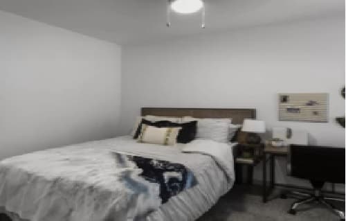 A cozy bedroom at The Hudson in College Station features a neatly made bed with white bedding, decorative pillows, a nightstand with a lamp, and a small desk with a chair against a white wall. The room is softly lit.