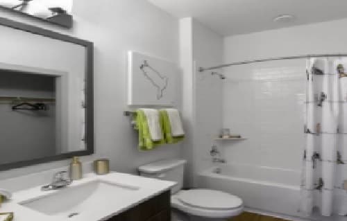 A modern bathroom in Hudson with a sink, mirror, toilet, and a bathtub with a shower curtain. Towels hang on a rack above the toilet, and simple wall art decorates the white wall—perfect for College Station living.