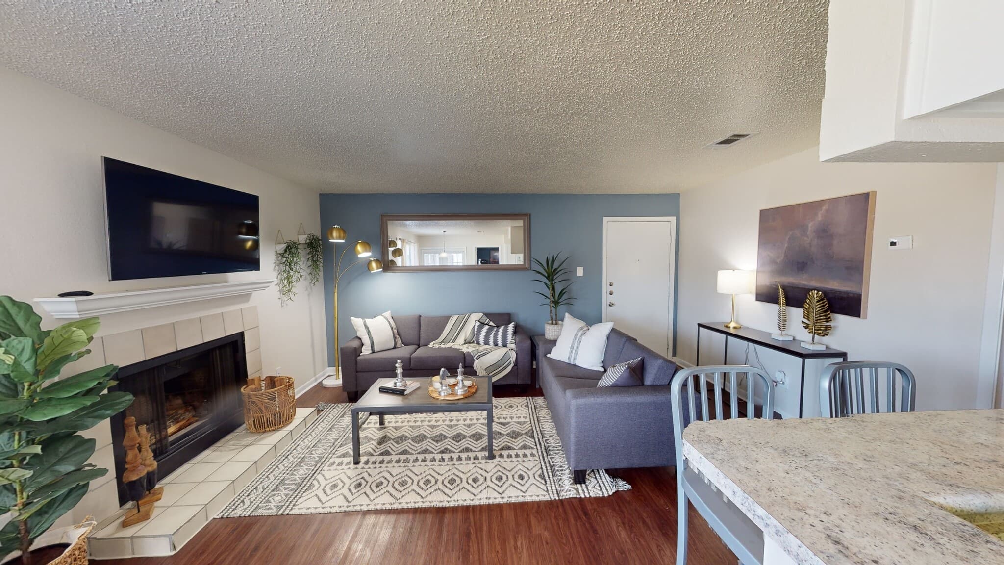 Modern living room at Landing College Station with a gray sectional sofa, patterned rug, and fireplace. A large wall mirror, mounted TV, decorative plants, and soft lighting create a cozy vibe that opens to a breakfast bar area.