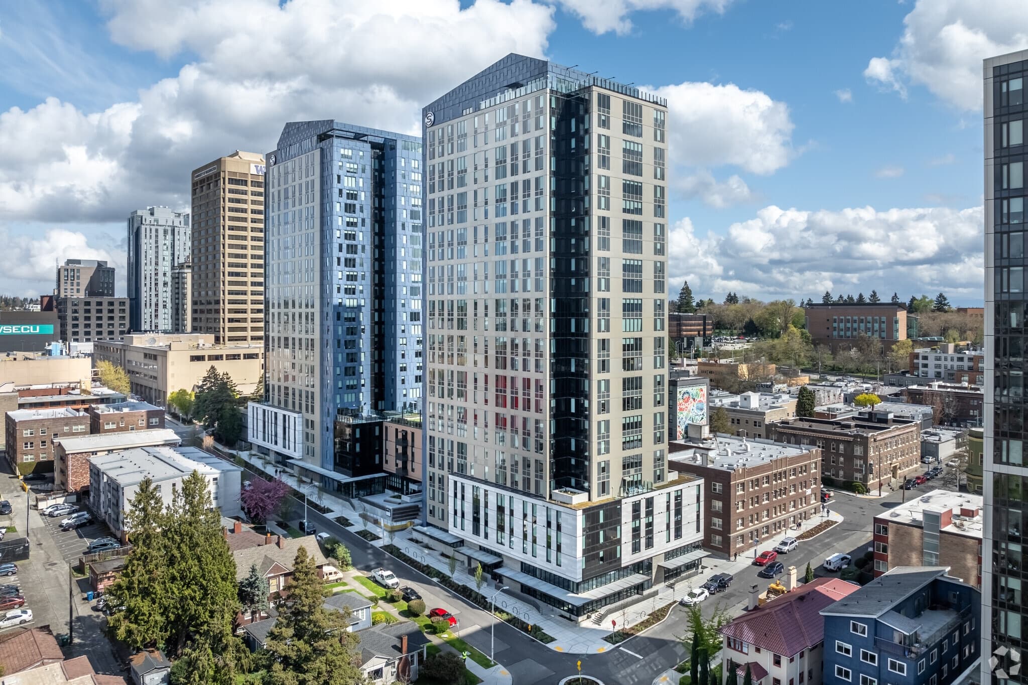 Aerial view of tall modern apartment buildings like The Standard at Seattle in an urban area, surrounded by smaller residential and commercial structures, with trees and a partly cloudy sky in the background.