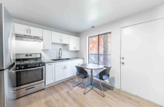 A modern, small kitchen at Elevation at the U Apartments Reno features white cabinets, stainless steel appliances, a sink, and a cozy dining table for two by a large window and white door. Light wood flooring brightens the space.