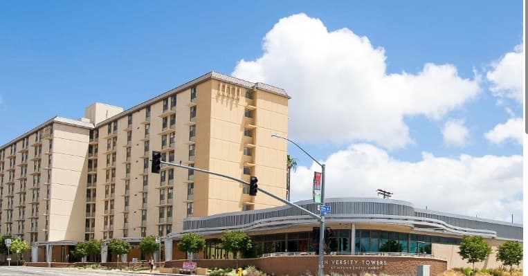 A tall beige building marked University Towers rises beside a modern, glass-fronted entrance under a blue sky with fluffy clouds. Trees and a streetlight add charm to the scene in front of University Towers.