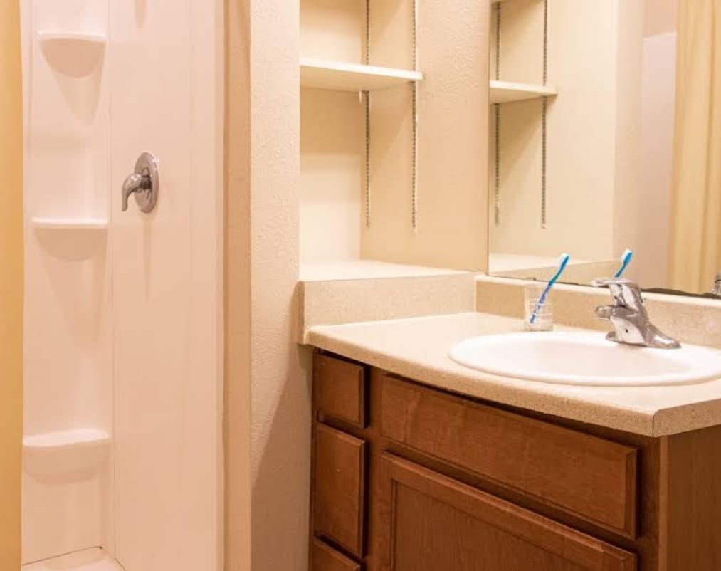 A bathroom at White Creek Apartments features a wooden cabinet, a cream countertop with sink, two blue toothbrushes in a holder, wall mirror, built-in shelving, and a white shower with built-in shelves and silver faucet.