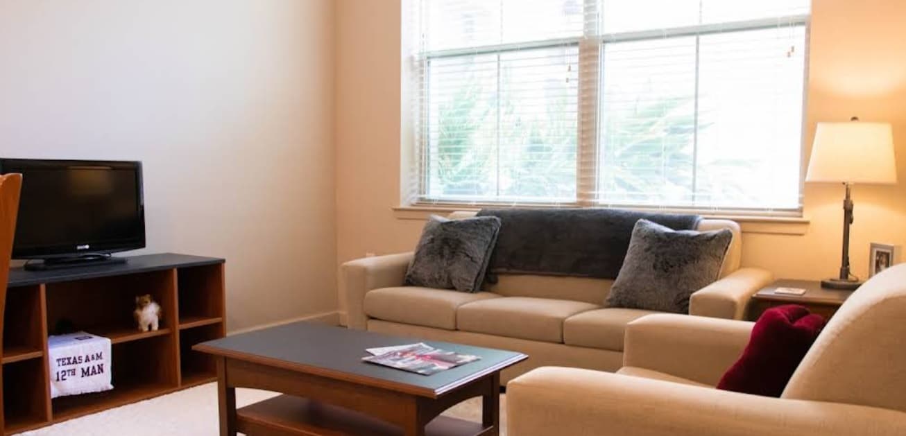 A cozy living room at White Creek Apartments features a beige sofa and armchair, a wooden coffee table with magazines, a TV on a stand, a lamp, and a large window letting in natural light.