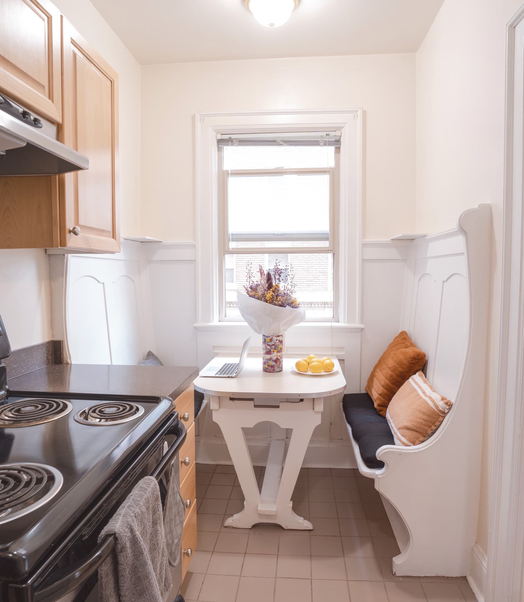 A small, cozy kitchen nook at Yugo Seattle features a white table with a vase of dried flowers, a bowl of lemons, and seating with black and orange cushions under a window. The Wilsonian charm is enhanced by bright natural light.