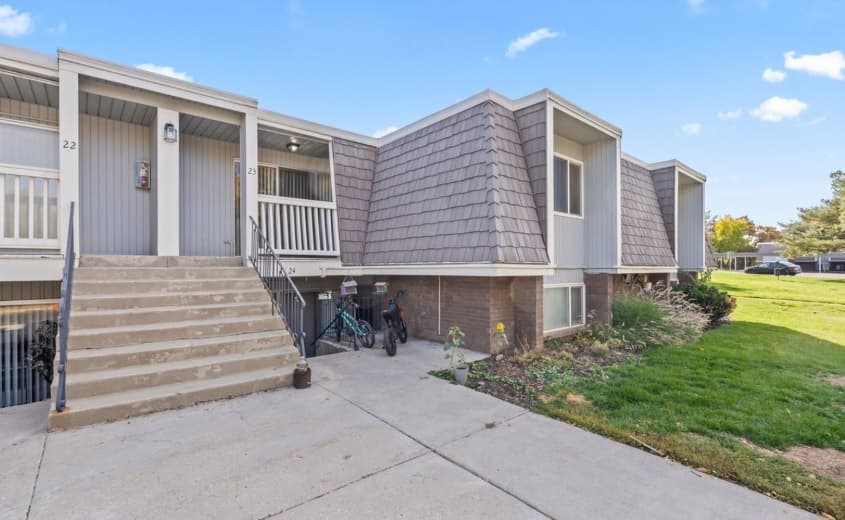 A two-story apartment building at Christopher Village Ogden features a shingled exterior, outdoor stairs to the upper unit, bicycles parked below, landscaped grass along the side, and a clear, sunny sky in the background.