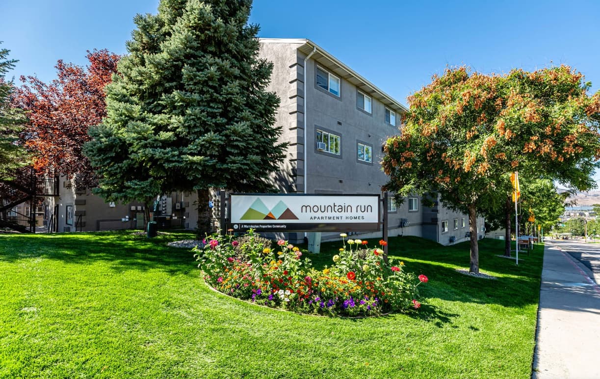 Mountain Run Apartment Homes Orem features a welcoming sign in front, surrounded by green grass, colorful flowers, and trees, creating a vibrant scene on a sunny day with blue sky.