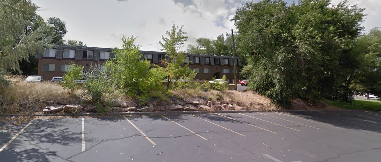 A mostly empty asphalt parking lot with faded lines sits in the foreground. Behind it, a grassy slope with rocks and trees leads up to Brownstone Apartments Ogden, where several cars are parked near the two-story brown building.