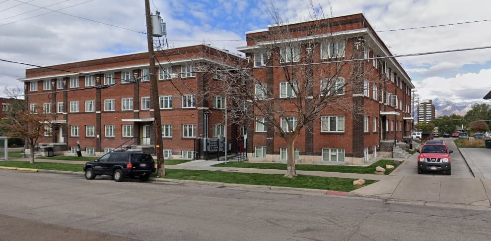 The McGregor Apartments Ogden is a three-story red brick apartment building with white trim on a corner, parked cars lining the street and bare trees in front. The sky above the McGregor Apartments is partly cloudy.