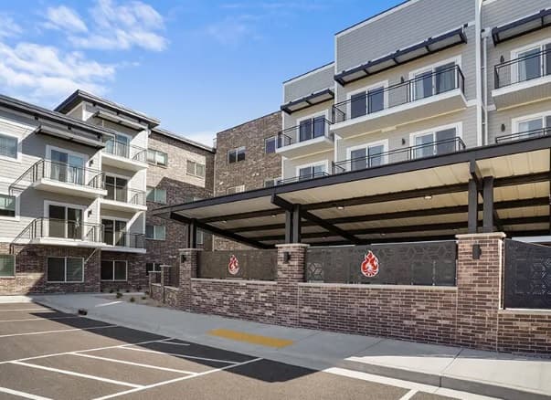 Phoenix Rise Ogden is a modern apartment building with balconies, brick and siding exterior, a covered parking area, and an empty parking lot in front under a partly cloudy sky.