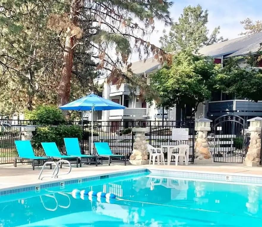 A clear outdoor swimming pool with blue lounge chairs, a blue umbrella, and a white table with chairs, surrounded by trees and residential buildings at Eagle Pointe Apartments, an Eagle Pointe Partners Property in Boise.